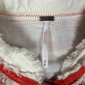 May include: Close-up of a cream-colored top with a fringed collar and red trim. The top features a tag that reads "Made in India / Fabriqu&eacute; en Inde" and the size "M". The collar is stitched with red thread.