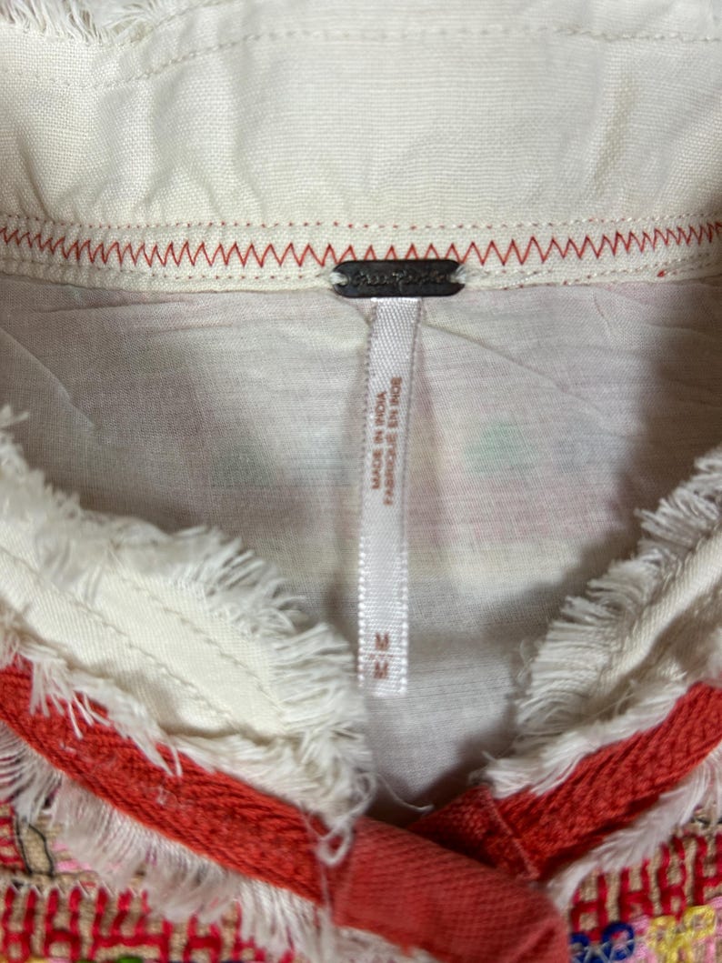 May include: Close-up of a cream-colored top with a fringed collar and red trim. The top features a tag that reads "Made in India / Fabriqu&eacute; en Inde" and the size "M". The collar is stitched with red thread.