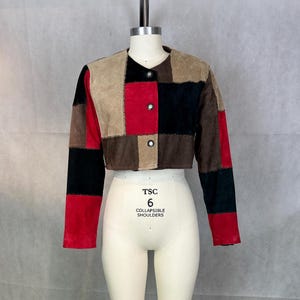 May include: A cropped jacket with a patchwork design in red, black, brown, and tan suede. The jacket has long sleeves and a round neckline, fastened with three silver buttons. The garment is displayed on a mannequin with the text "TSC 6 COLLAPSIBLE SHOULDERS".