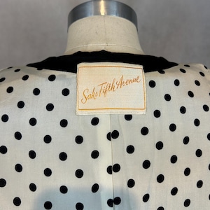 Puede incluir: Vista posterior de un vestido de lunares blancos y negros con cuello de terciopelo negro. Una etiqueta blanca con letras doradas dice "Saks Fifth Avenue".