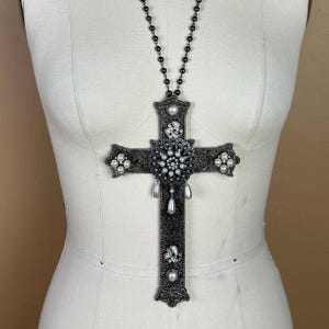 Può includere: Una collana con pendente a croce grande e ornata. La croce in metallo scuro è impreziosita da perle, cristalli e filigrana decorativa. La collana presenta una catena di perline e pendenti a forma di pera. La croce è esposta su un manichino.