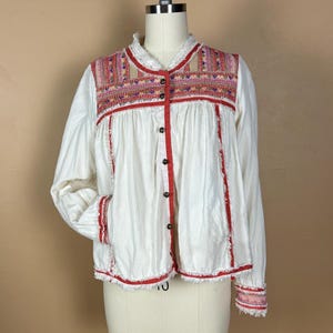 May include: White button-down blouse with red trim and embroidered details. The blouse features a mandarin collar and long sleeves. The yoke and cuffs have colorful embroidered patterns. The blouse is displayed on a mannequin.