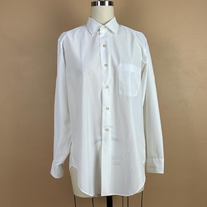 May include: A white long-sleeve shirt with a button-down front, a chest pocket, and a classic collar. The shirt is displayed on a mannequin. The buttons are gold-colored. The shirt has a curved hemline.