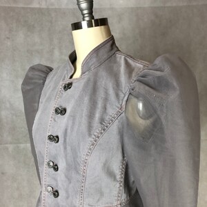 K&ouml;nnte beinhalten: Eine graue Jeansjacke mit Stehkragen und Puff&auml;rmeln. Die Jacke hat eine Reihe schwarzer Kn&ouml;pfe auf der Vorderseite.