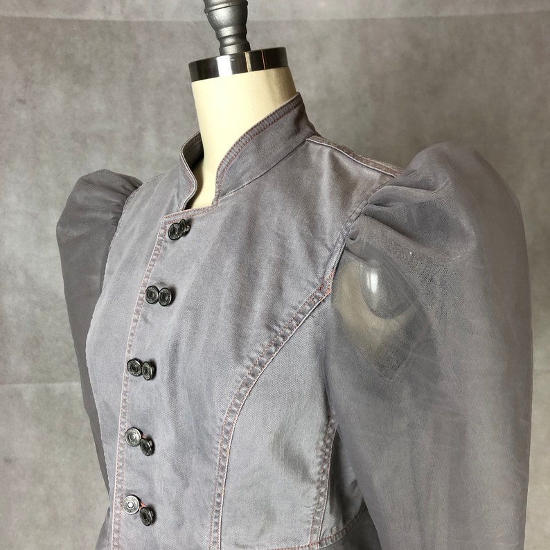K&ouml;nnte beinhalten: Eine graue Jeansjacke mit Stehkragen und Puff&auml;rmeln. Die Jacke hat eine Reihe schwarzer Kn&ouml;pfe auf der Vorderseite.