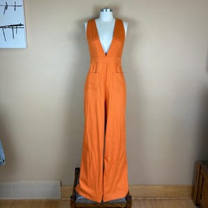 May include: A sleeveless, vibrant orange jumpsuit featuring a deep V-neckline, a defined waistline, and two front patch pockets. The garment has wide-leg trousers that extend to the floor, creating a long, continuous silhouette in a solid colour.