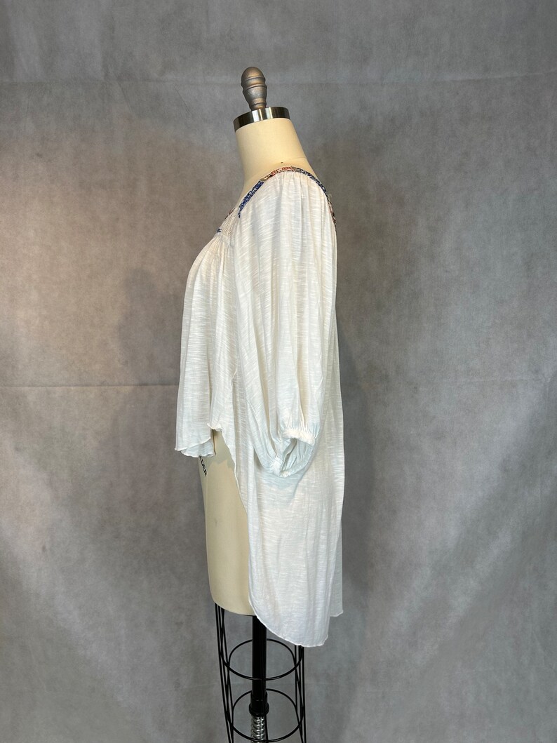 Puede incluir: Blusa blanca, ligera y semitransparente, con un dobladillo alto-bajo y detalles bordados alrededor del escote. La blusa tiene un ajuste holgado y mangas 3/4.