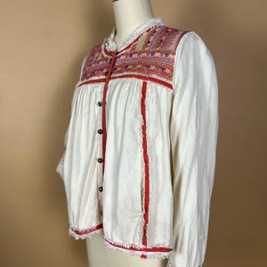 May include: White long-sleeved jacket with colorful embroidered yoke and red trim. The jacket features a ruffled collar, button-down front, and frayed edges. The cuffs also have embroidered details. The garment is displayed on a mannequin.