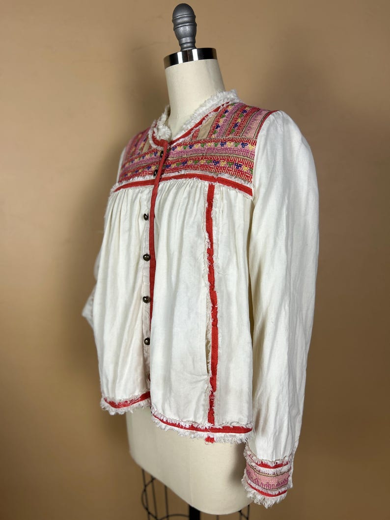 May include: White long-sleeved jacket with colorful embroidered yoke and red trim. The jacket features a ruffled collar, button-down front, and frayed edges. The cuffs also have embroidered details. The garment is displayed on a mannequin.