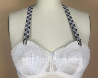 May include: White strapless bra with lace detailing and a small blue button. The bra has removable straps with a black and white checkered pattern. The bra is displayed on a mannequin.