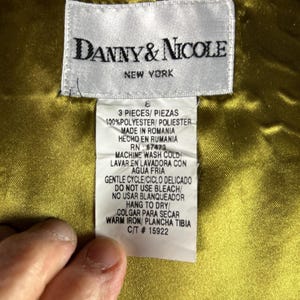 May include: A tag from Danny & Nicole, New York, with care instructions. The tag is white with black text and includes washing instructions, material composition, and the text "3 PIECES/PIEZAS". The background is a golden-yellow fabric.