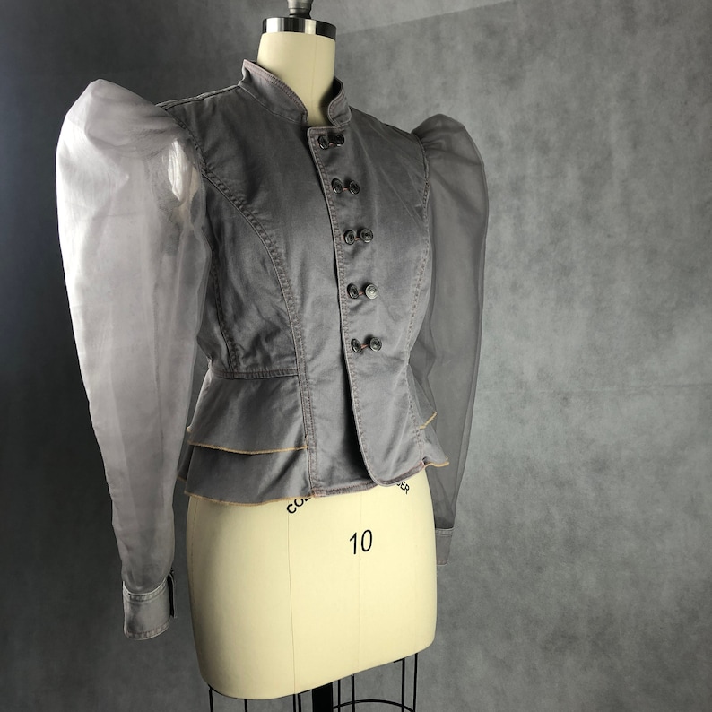 K&ouml;nnte beinhalten: Eine graue Jeansjacke mit einem R&uuml;schen-Saum und durchsichtigen, bauschigen &Auml;rmeln. Die Jacke hat einen Stehkragen und einen Knopfverschluss.