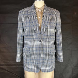 May include: A blue, black, and white plaid blazer with a single-button closure. The blazer has two front pockets and is displayed on a mannequin. The blazer is paired with a tan collared shirt.