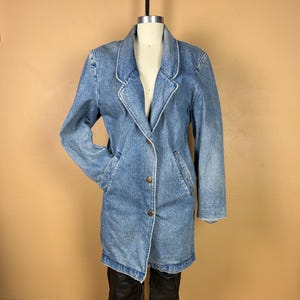 May include: A light blue denim blazer with a classic collar and button closure. The long-sleeved jacket features two front pockets and a tailored fit. The blazer is displayed on a mannequin.