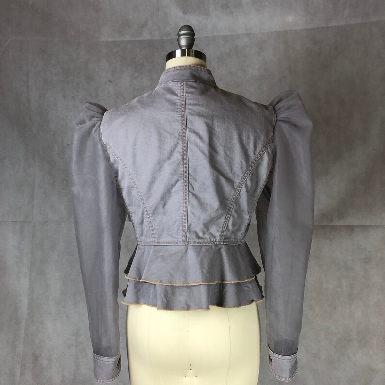 K&ouml;nnte beinhalten: Eine graue, kurze Jacke mit gerafftem Saum und langen &Auml;rmeln. Die Jacke hat eine figurbetonte Silhouette und einen Stehkragen.