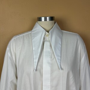 May include: A white long-sleeve button-up shirt featuring a prominent, oversized pointed collar. The shirt is constructed with a vertical pleated front panel design, a single visible top button, and long sleeves, displayed on a mannequin torso.