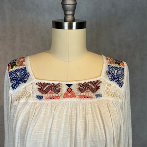 Puede incluir: Una blusa blanca de algod&oacute;n con cuello cuadrado y patrones geom&eacute;tricos bordados en azul, naranja y rojo.