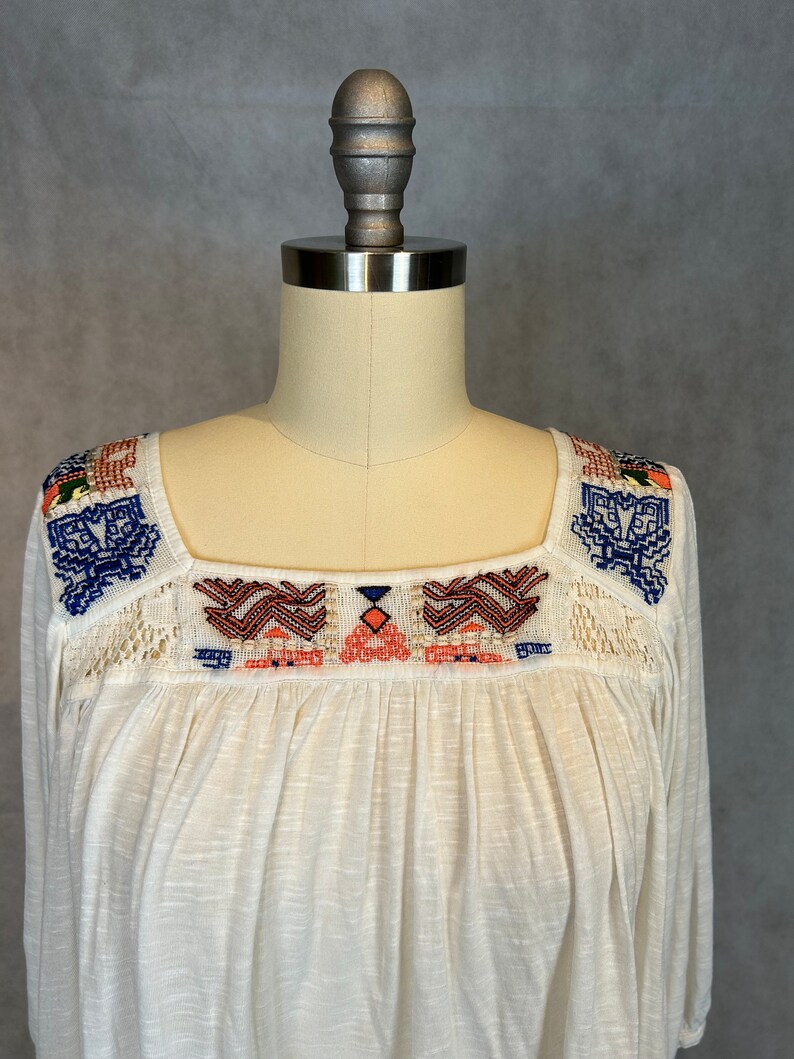 Puede incluir: Una blusa blanca de algod&oacute;n con cuello cuadrado y patrones geom&eacute;tricos bordados en azul, naranja y rojo.