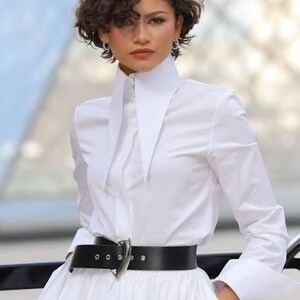 May include: A crisp white long-sleeved button-down shirt featuring an oversized, pointed collar and wide cuffs. The shirt is tucked into a wide black leather belt with a prominent silver-toned metal buckle, cinching the waist of a full white skirt.