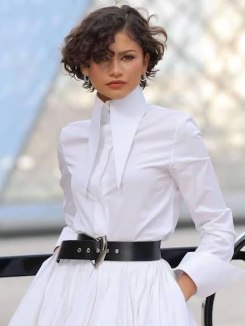 May include: A crisp white long-sleeved button-down shirt featuring an oversized, pointed collar and wide cuffs. The shirt is tucked into a wide black leather belt with a prominent silver-toned metal buckle, cinching the waist of a full white skirt.