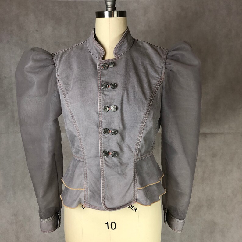 K&ouml;nnte beinhalten: Eine graue Jeansjacke mit gerafftem Saum und Puff&auml;rmeln. Die Jacke hat einen Stehkragen und einen Knopfverschluss.
