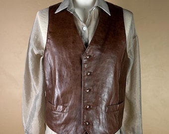 May include: A brown leather vest with a button-up front and two front pockets. The vest is worn over a light-colored collared shirt. The vest appears to be made of genuine leather and has a slightly textured surface. The overall style is classic and timeless.