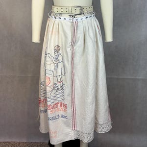 May include: A white cotton skirt with a red and white stripe down the side. The skirt has a lace trim at the bottom and features an embroidered image of a woman pouring sugar from a bag. The text on the bag reads "Satin Brand Sugar Mills Inc."