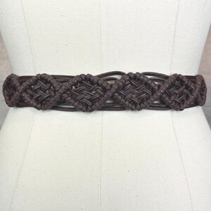 May include: A brown macrame belt with a woven design. The belt features a series of diamond-shaped woven patterns. The belt is displayed on a mannequin.