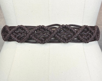 Puede incluir: Un cinturón de macramé marrón con un diseño tejido. El cinturón presenta una serie de patrones tejidos en forma de diamante. El cinturón se muestra en un maniquí.