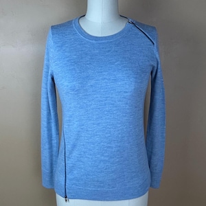 May include: A light blue long-sleeve sweater featuring a round neckline and a diagonal zipper detail that extends from the left shoulder down to the bottom hem. The garment is crafted from a fine-knit fabric with a subtle heathered texture throughout.