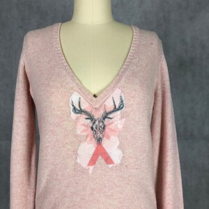 Könnte beinhalten: Ein rosa V-Ausschnitt-Pullover mit einem grafischen Druck eines Hirschschädels mit Geweih und geometrischen Formen.