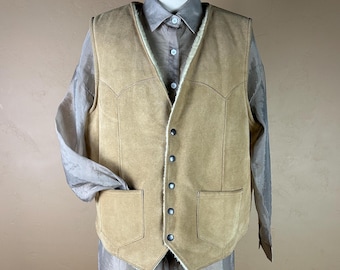 May include: A tan suede vest with a cream-colored shearling lining and two front pockets. The vest has a button-up closure and decorative stitching along the edges. The vest is displayed on a mannequin over a light brown button-down shirt.