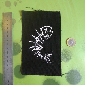 K&ouml;nnte beinhalten: Ein schwarzer Stoff-Patch mit einem wei&szlig;en gestickten Bild eines Fischskeletts mit einem Totenkopf und gekreuzten Knochen auf dem Kopf.