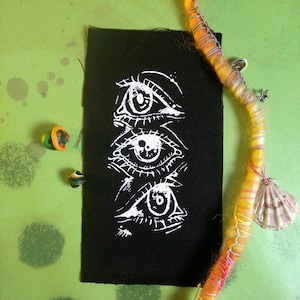 May include: A black fabric patch featuring a white illustration of three stylized eyes stacked vertically. The patch is set against a green background with colorful beads and a decorative strand with a seashell and star charm.