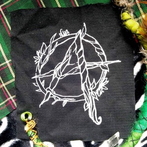 Könnte beinhalten: Ein weiß gestickter Aufnäher auf schwarzem Stoff mit einem stilisierten Anarchiesymbol, umgeben von einem Kreis aus Dornen.