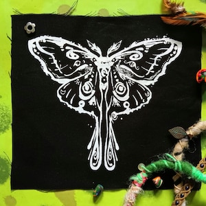 May include: A black square fabric patch featuring a detailed white moth design. The moth has intricate wing patterns and a symmetrical body. The patch is secured with a silver-colored flower-shaped pin. The background is a bright green with decorative elements.