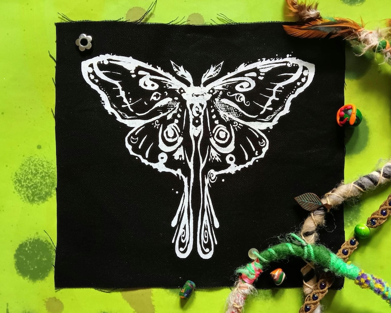 May include: A black fabric patch with a white moth design. The moth's wings and body have detailed patterns. The patch is pinned with a silver flower-shaped pin. The background is a bright green with decorative elements. The patch is approximately 20 cm square.