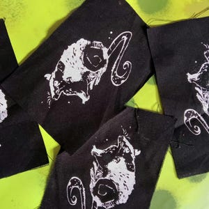 Op de afbeelding: Verschillende zwarte stoffen patches met een witte illustratie van een gehoornd wezen. Het wezen heeft een gestileerd gezicht en een gebogen, decoratief element. De patches zijn gerangschikt op een felgroene achtergrond.