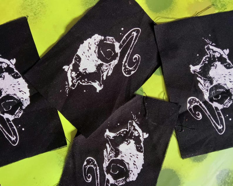 Op de afbeelding: Verschillende zwarte stoffen patches met een witte illustratie van een gehoornd wezen. Het wezen heeft een gestileerd gezicht en een gebogen, decoratief element. De patches zijn gerangschikt op een felgroene achtergrond.
