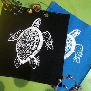Puede incluir: Dos parches de tela con un diseño de tortuga marina blanca. La tortuga en el parche negro tiene detalles intrincados, mientras que la tortuga en el parche azul tiene un diseño más simple.