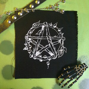 May include: A black patch featuring a white pentagram design. The pentagram is encircled by a circle of twigs, leaves, and beads.