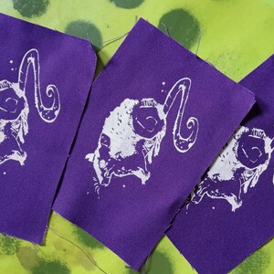 Op de afbeelding: Drie paarse stoffen patches met een wit ram schedel ontwerp. De ram schedel heeft grote, gebogen hoorns en is gecentreerd op de patches. De patches zijn gerangschikt op een groen en gele achtergrond.