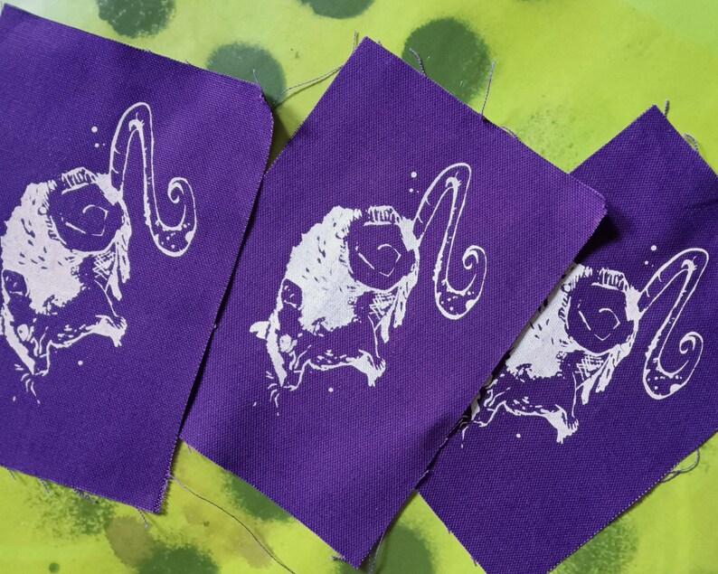 Op de afbeelding: Drie paarse stoffen patches met een wit ram schedel ontwerp. De ram schedel heeft grote, gebogen hoorns en is gecentreerd op de patches. De patches zijn gerangschikt op een groen en gele achtergrond.