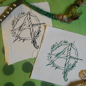 Könnte beinhalten: Zwei weiße Stoffpatches mit schwarzen und grünen Stickereien. Die Designs zeigen einen stilisierten Buchstaben A in einem Kreis aus Blättern und Ranken.