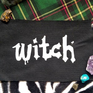 Könnte beinhalten: Schwarzer Stoff-Patch mit dem Wort "witch" in weißer, tropfender Schrift.