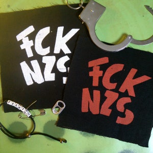 Könnte beinhalten: Zwei schwarze Stoff-Patches mit dem Schriftzug "FCK NZS" in weißer und roter Farbe. Die Patches liegen auf einer grünen Oberfläche mit Handschellen und einem Armband mit dem Schriftzug "FXXK YOU".