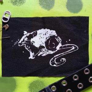 Pode incluir: Patch de tecido preto com uma ilustração branca de um rato. O rato é retratado de forma estilizada e artística. Um alfinete de segurança prateado, um puxador prateado e um cinto preto com ilhós prateados também são visíveis.