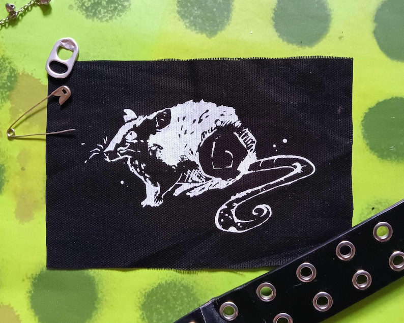 Op de afbeelding: Zwarte stoffen patch met een witte illustratie van een rat. De rat is afgebeeld op een gestileerde, artistieke manier. Een zilveren veiligheidsspeld, een zilveren treklipje en een zwarte riem met zilveren oogjes zijn ook zichtbaar.