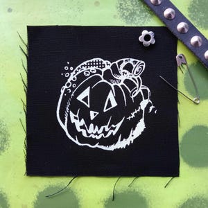 Pode incluir: Um remendo de tecido preto com um desenho branco de uma abóbora de Halloween com a boca costurada e um pequeno desenho detalhado no topo da cabeça.