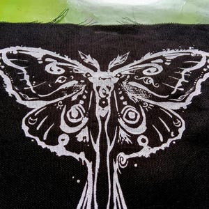 May include: Black fabric patch with a white moth design. The moth's wings and body are intricately detailed. The white design stands out against the black background, creating a bold contrast.